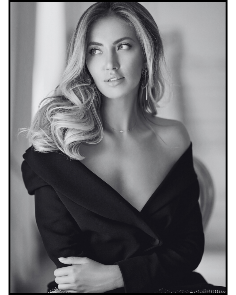 A black and white photo of a white woman with blonde hair wearing a black off the shoulder dress with a deep v at the neckline. Her hair lays in waves over one shoulder